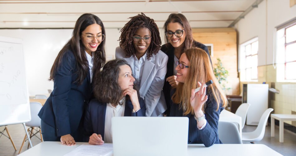 Content businesswomen working with laptop. Front view of cheerful multiethnic female colleagues using laptop computer and discussing work in office. Women in business concept
